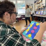 Two women in a classroom, one seated and holding a colorful geometric artwork, the other standing near displayed pieces.